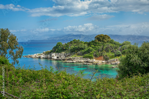 Beautiful summer landscape of sea lagoon with turquoise calm water, cliffs and rocks on the shore, meadow with green grass, trees and bushes, mountains on the horizon. Corfu Island, Greece