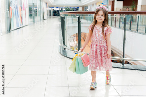 Portrait of a little happy girl in the mall. A smiling laughing girl in a pink dress with multi-colored bags in her hands is walking around the mall, looking at the shop windows