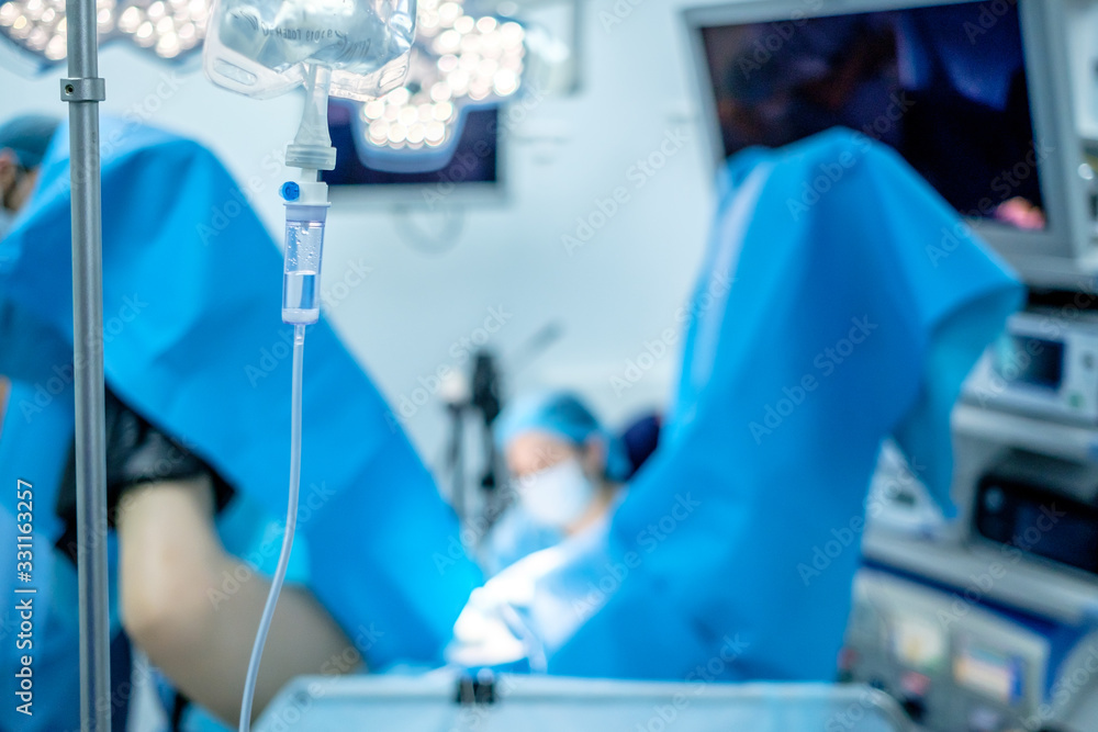 Obraz premium Transparent plastic tubes and dropper for a medical dropper with saline solution. Against a very blurry background, surgeons perform a surgical operation in the light of bright lamps.