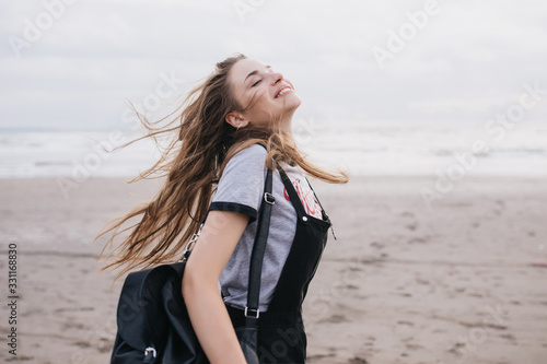Romantic long-haired woman with black backpack enjoying good day at sandy beach. Outdoor shot of caucasian lovely girl jumping on sky background.