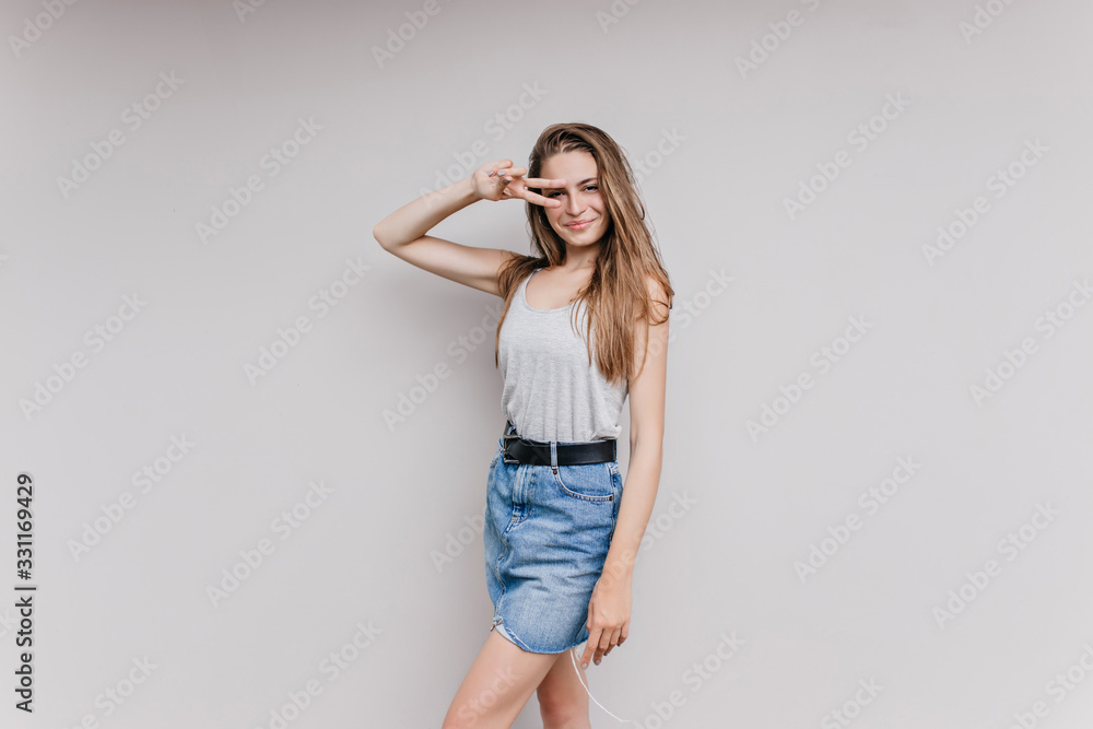 Pretty smiling girl in denim skirt posing with peace sign in studio. Indoor portrait of wonderful caucasian lady having fun during photoshoot. Pretty smiling girl in denim skirt posing with peace sign in studio. Indoor portrait of wonderful caucasian lady having fun during photoshoot.