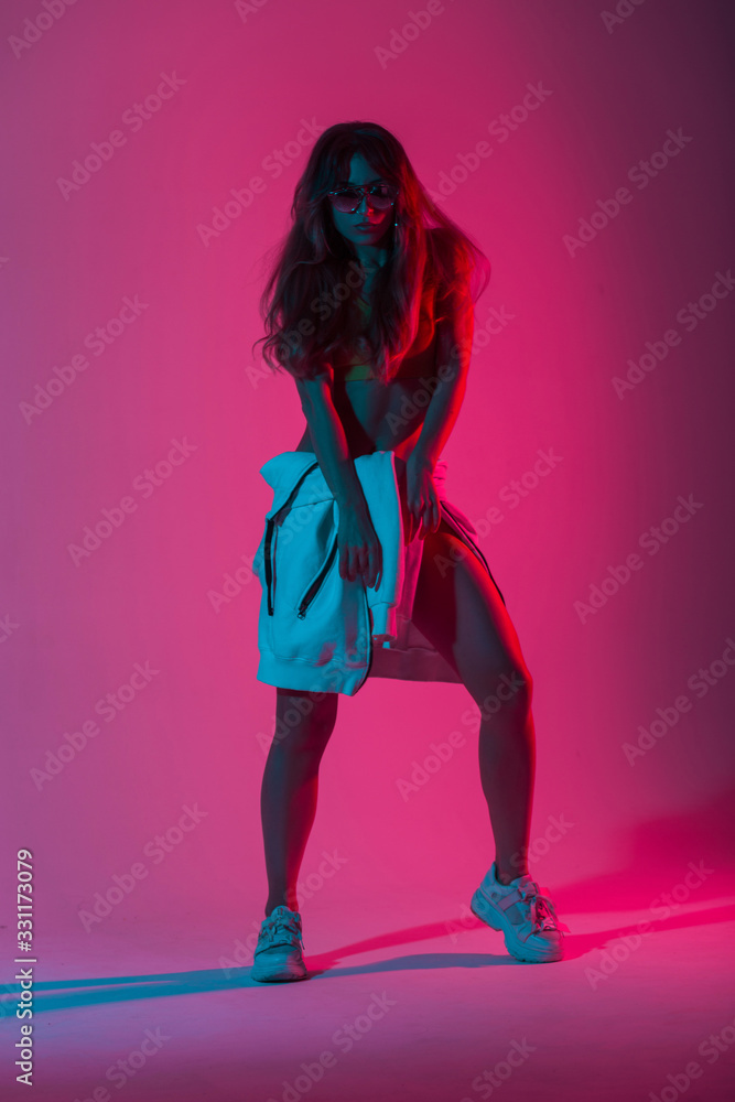 Fototapeta Young sexy woman in a fashion top in hoodie in shoes posing in a studio with bright neon pink light. Girl hipster in sunglasses in trendy wear stands indoors with a multi-colored color in style disco.
