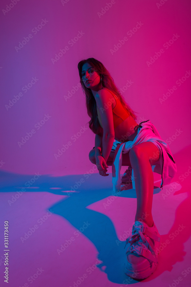 Fototapeta Sporty pretty young woman dancer with stylish hoodie in a top in sneakers sits in a room with bright blue-violet light.Sexy girl model posing in trendy wear indoors with neon pink color in disco style
