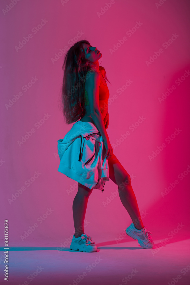 Fototapeta Pretty sexy cool young woman in fashionable sportswear with a vintage hoodie posing indoors with multicolored neon color. Stylish attractive slim sexy girl in the studio with bright pink disco light.