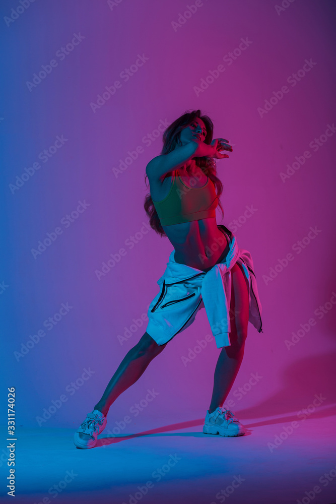 Fototapeta Young woman dancer in stylish youth clothes in gym shoes dancing and posing in a room with bright neon blue-pink color. Sports girl enjoys a dance in the studio with multi-colored ultraviolet light.