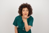 Cute appealing doctor female wearing medical uniform blows kiss at camera, demonstrates love to boyfriend or says goodbye on distance, isolated over studio background.