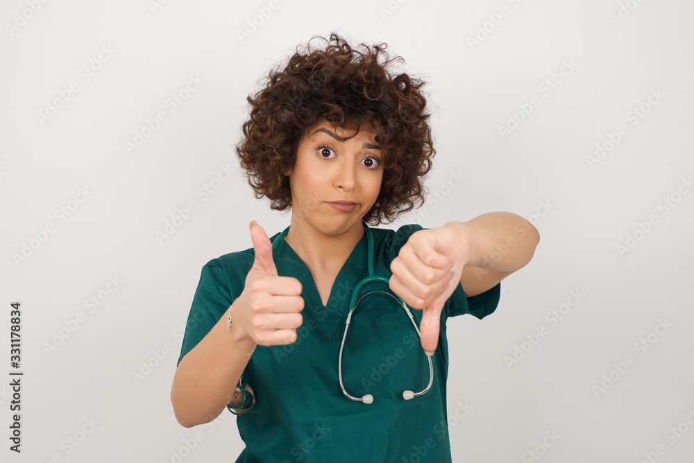 Pretty young pensive doctor woman making good-bad sign. Displeased and unimpressed wearing green medical uniform and standing against gray wall. Pretty young pensive doctor woman making good-bad sign. Displeased and unimpressed wearing green medical uniform and standing against gray wall.
