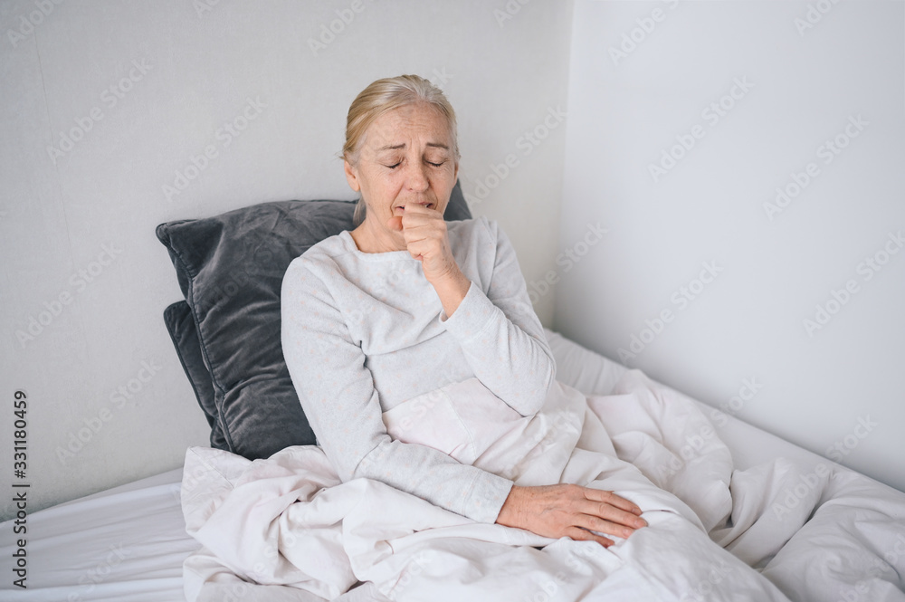 Senior woman coughing in fist sick of coronavirus viral infection spreading corona virus covering mouth and nose. Painful cough ill elderly patient lying in bed at home quarantine. First symptoms Senior woman coughing in fist sick of coronavirus viral infection spreading corona virus covering mouth and nose. Painful cough ill elderly patient lying in bed at home quarantine. First symptoms