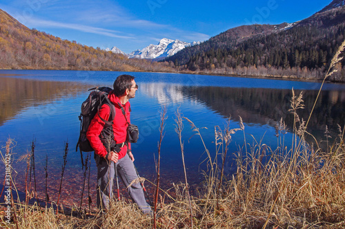 A middle-aged man in a red jacket and glasses with a backpack for tourism on the background of the lake which reflects the snow-capped mountains.