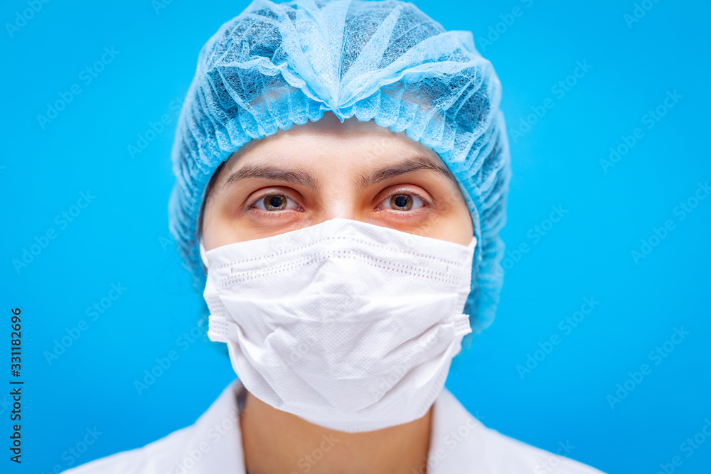 Young female doctor in medicine mask, hair cap and white gown portaiture on a blue background closeup shot. Shallow depth of field. Coronavirus, pandemia and medical care concept. Young female doctor in medicine mask, hair cap and white gown portaiture on a blue background closeup shot. Shallow depth of field. Coronavirus, pandemia and medical care concept.