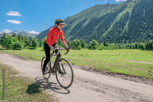 A young smiling girl on a cyclocross bike rides along a winding mountain road against a background of green forest and mountains with glaciers and snow on the tops