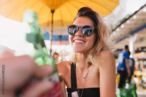 Gorgeous tanned girl in black sunglasses spending summer day in outdoor restaurant. Laughing blonde woman wears golden accessories smiling during rest in cafe.