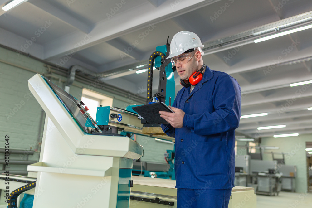 CNC machine operator in the Hard white Hat Walks Through Light Modern Factory While Holding tablet. Successful, Handsome Man in Modern Industrial Environment. CNC machine operator in the Hard white Hat Walks Through Light Modern Factory While Holding tablet. Successful, Handsome Man in Modern Industrial Environment.