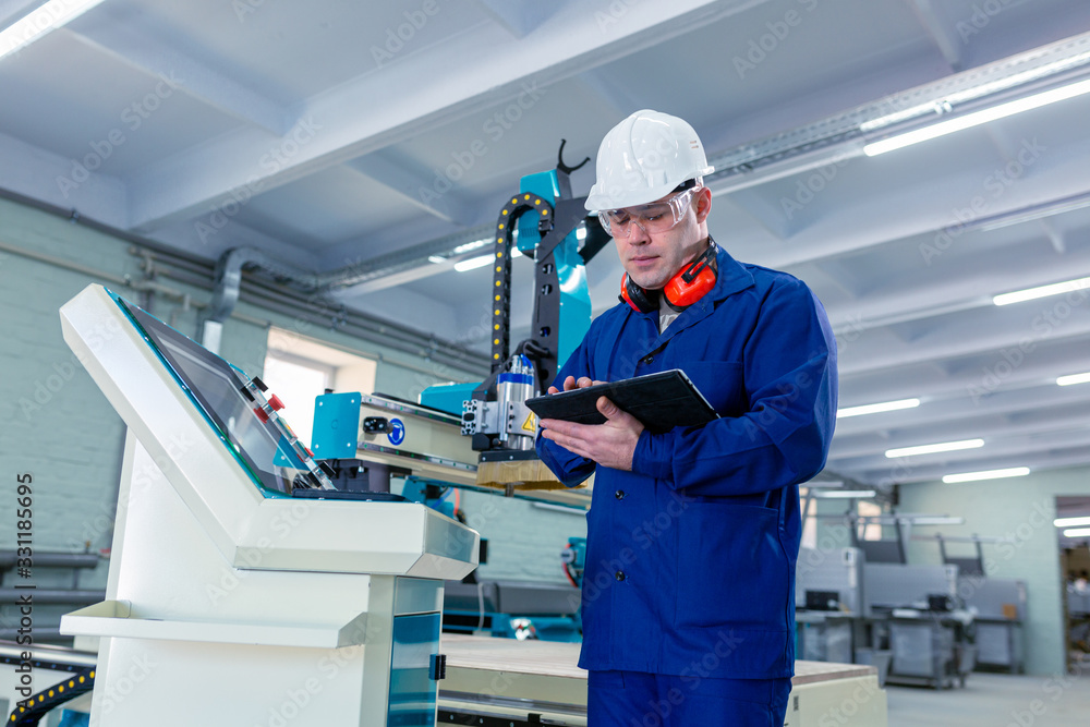 CNC machine operator in the Hard white Hat Walks Through Light Modern Factory While Holding tablet. Successful, Handsome Man in Modern Industrial Environment. CNC machine operator in the Hard white Hat Walks Through Light Modern Factory While Holding tablet. Successful, Handsome Man in Modern Industrial Environment.