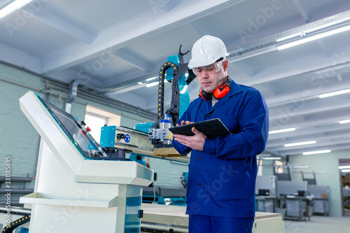 CNC machine operator in the Hard white Hat Walks Through Light Modern Factory While Holding tablet. Successful, Handsome Man in Modern Industrial Environment.