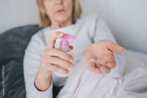 Hand sanitizer bottle senior woman using dispenser gel rub for corona virus hands hygiene coronavirus COVID-19 pandemic prevention. Patient lying in bed at home quarantine
