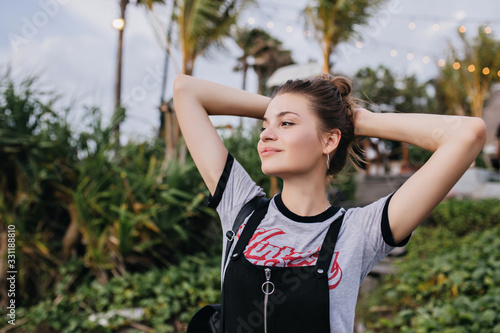 Blissful european girl looking away with romantic smile. Outdoor photo of dreamy young woman in trendy clothes posing with hands up on nature background.