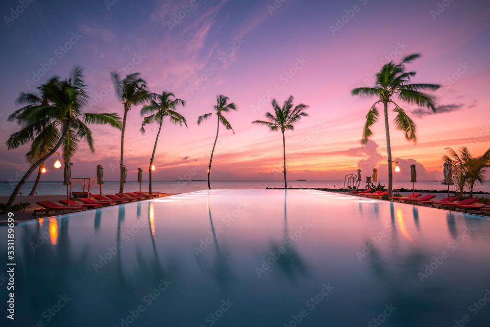 Luxury sunset over infinity pool in a summer beachfront hotel resort at tropical landscape. Tranquil beach holiday vacation background mood. Amazing island sunset beach view, palms swimming pool