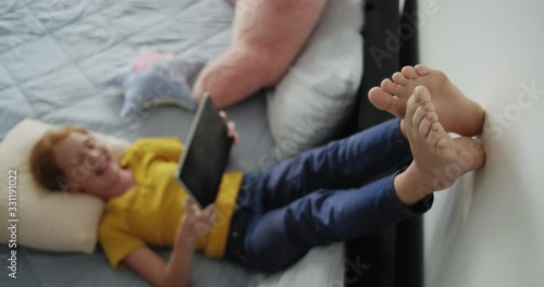Little girl watching movie in bedroom. Barefoot young people lying in bed at home. Happy redhead female child looking at video with laptop computer and smiling