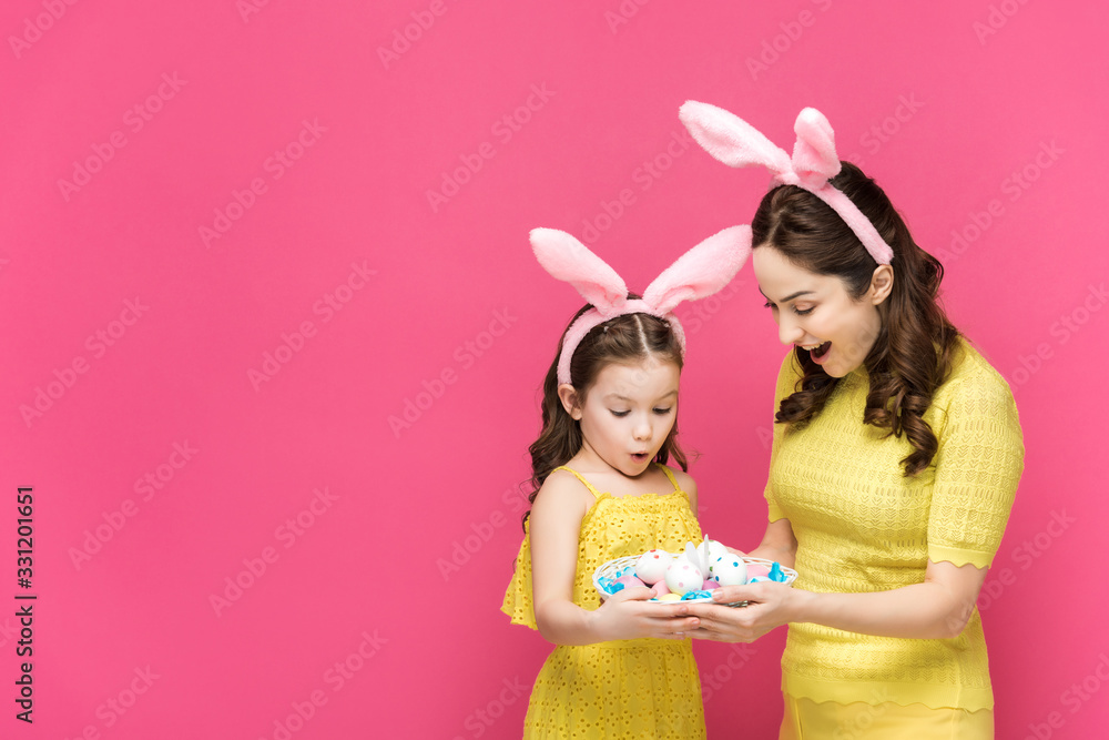 surprised mother and daughter in bunny ears looking at easter eggs isolated on pink