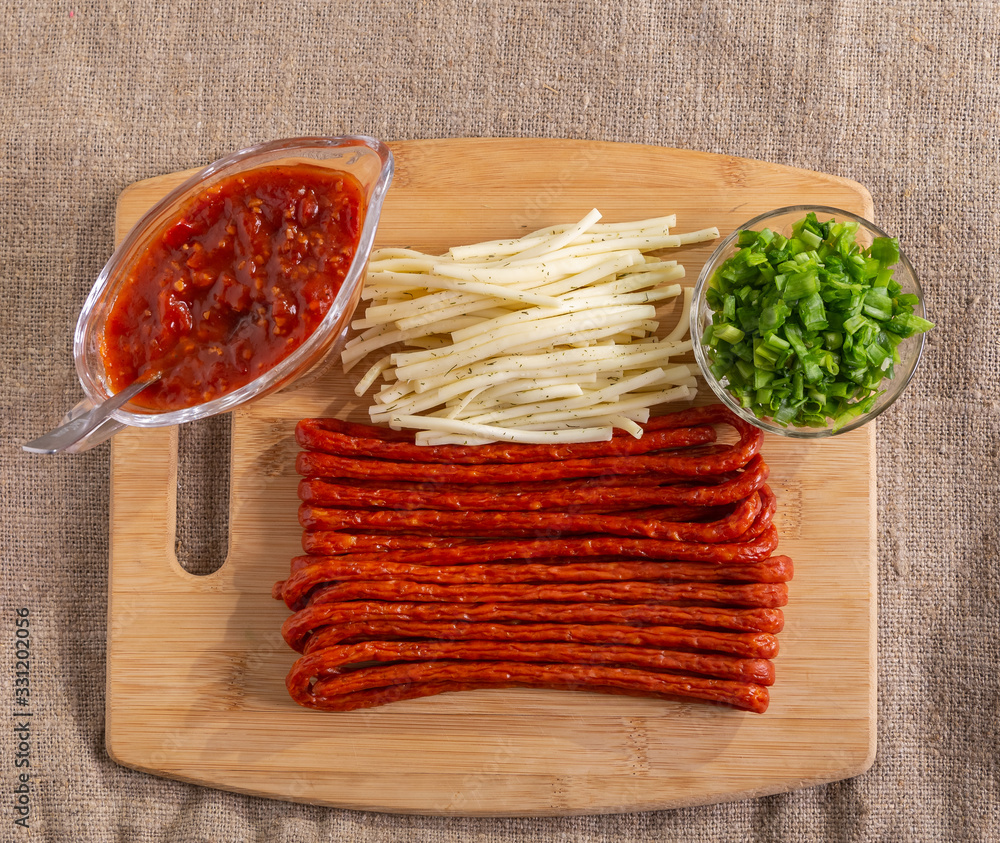 On the cutting Board are thin smoked sausages and cheese. Tomato sauce in a saucepan and chopped onions in a small plate stand next to each other . On the cutting Board are thin smoked sausages and cheese. Tomato sauce in a saucepan and chopped onions in a small plate stand next to each other .