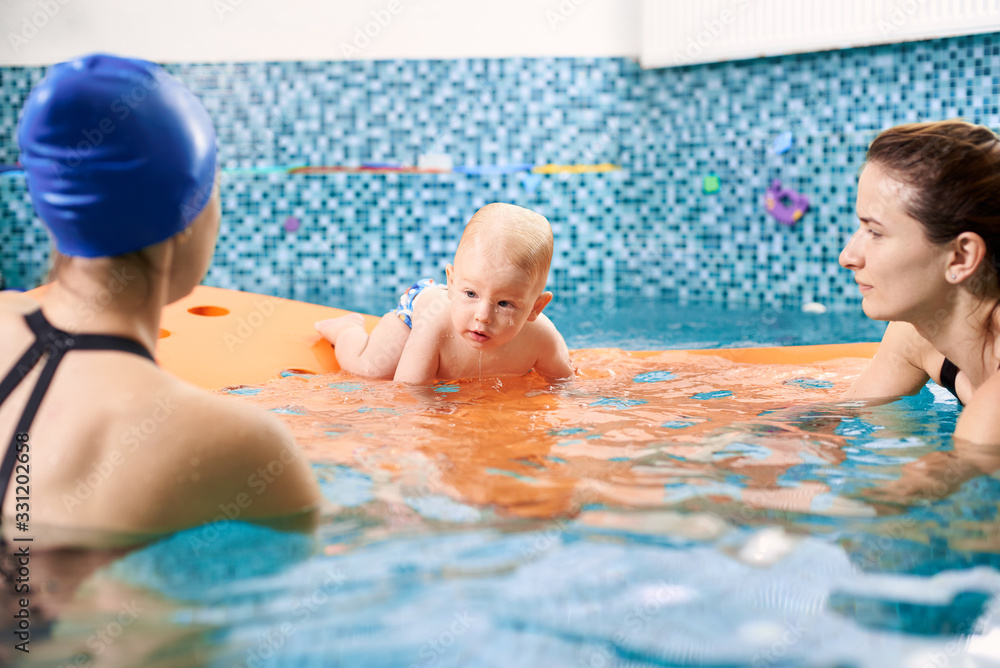 Wet concentrated little toddler is trying to crawl on a floating orange mat with holes, with the help of mother and instructor during swimming lesson in pool.