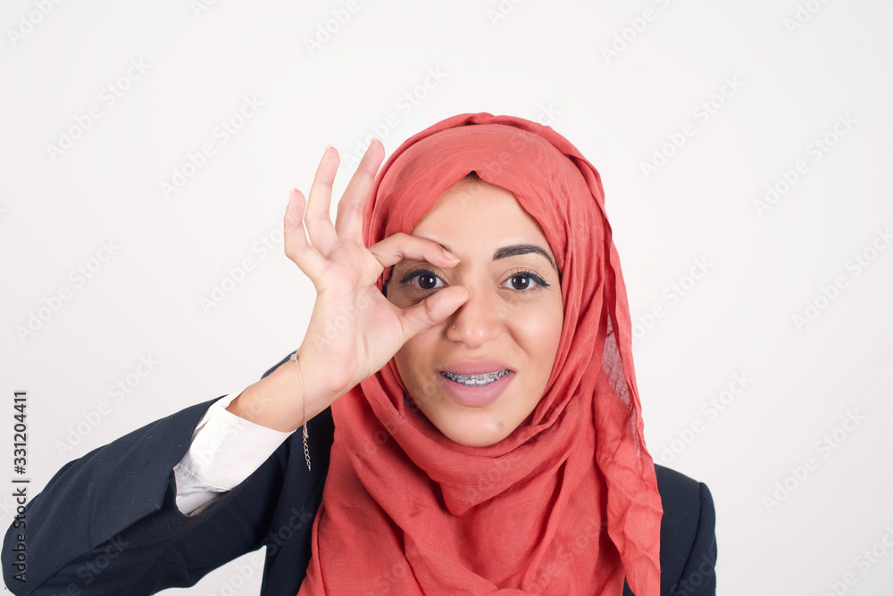 Beautiful young blonde woman with happy face smiling doing ok sign with hand on eye looking through finger. Wearing casual clothes and standing. Beautiful young blonde woman with happy face smiling doing ok sign with hand on eye looking through finger. Wearing casual clothes and standing.