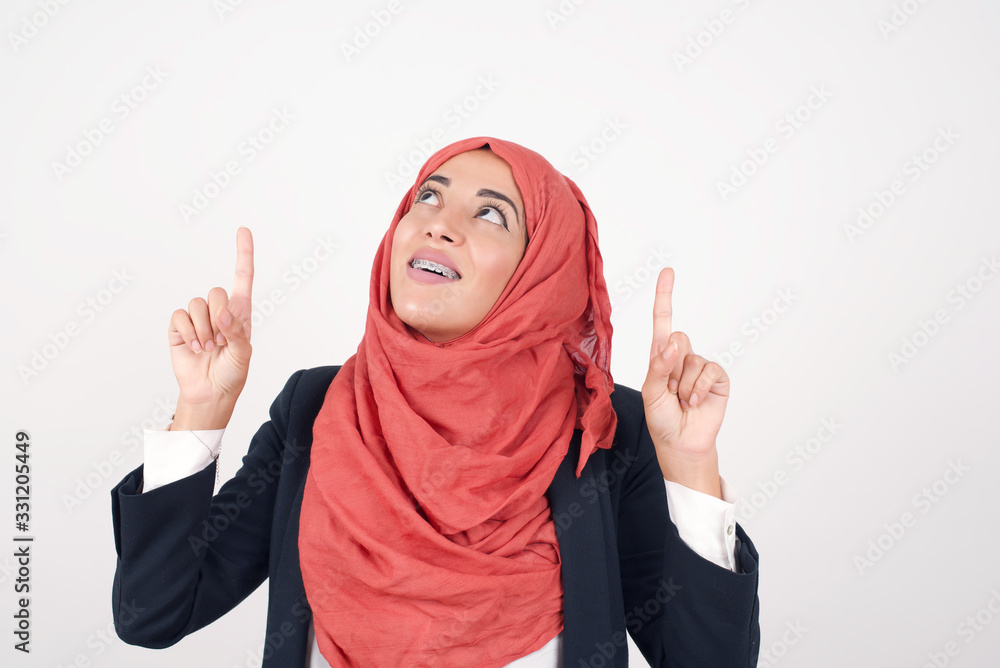 Horizontal shot of successful friendly looking young dark haired woman with bright make up exclaiming excitedly, pointing both index fingers up, indicating something shocking on blank copy space wall. Horizontal shot of successful friendly looking young dark haired woman with bright make up exclaiming excitedly, pointing both index fingers up, indicating something shocking on blank copy space wall.