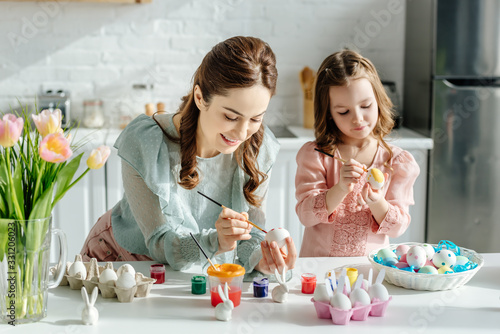 Selective focus of cute child and mother painting easter eggs near tulips