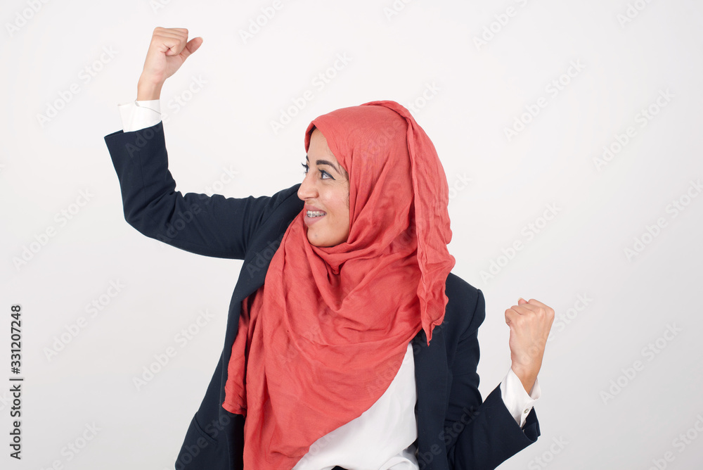 Attractive young muslim woman celebrating a victory punching the air with her fists and a beaming toothy smile over a blue studio background with copy space. Attractive young muslim woman celebrating a victory punching the air with her fists and a beaming toothy smile over a blue studio background with copy space.