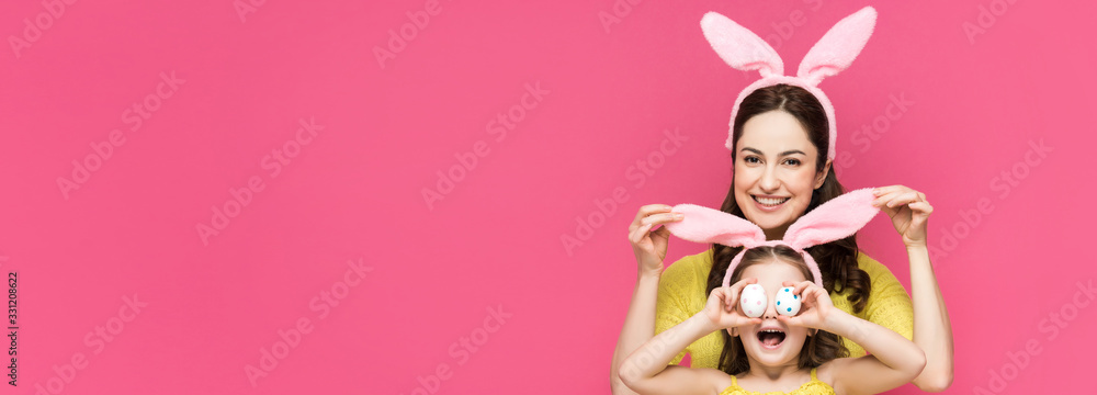 panoramic shot of happy mother touching bunny ears of daughter covering eyes with easter eggs isolated on pink
