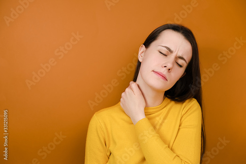 Displeased young woman feeling stiff sore neck pain concept rubbing massaging tensed muscles suffer from fibromyalgia, has gloomy expression, wears sweater, isolated on orange background