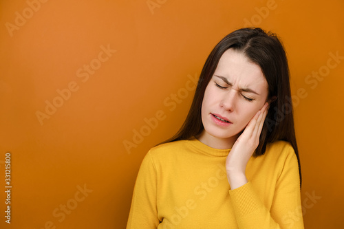 Tired young female suffering from strong earache. Stressed frowning girl plugging ear, feeling painful discomfort, avoids bad sound, wears yellow sweater, models against orange studio background.