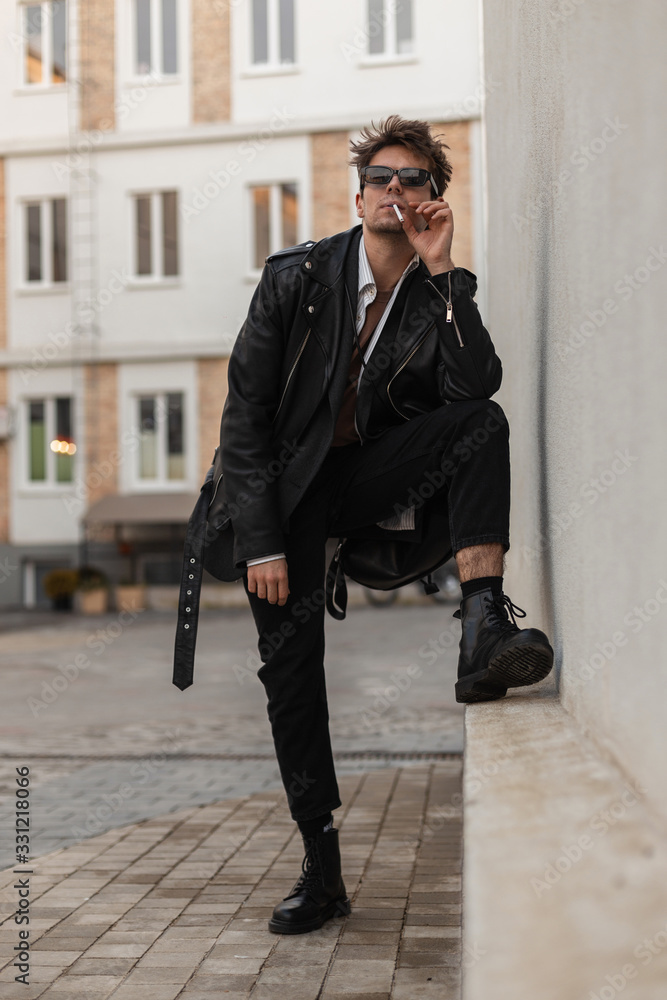 Young American man hipster in fashion black clothes in trendy sunglasses smokes near wall outdoors. Stylish guy model with cigarette enjoy nicotine. Pleasure of smoking. Relaxing but harmful habit. Ph...