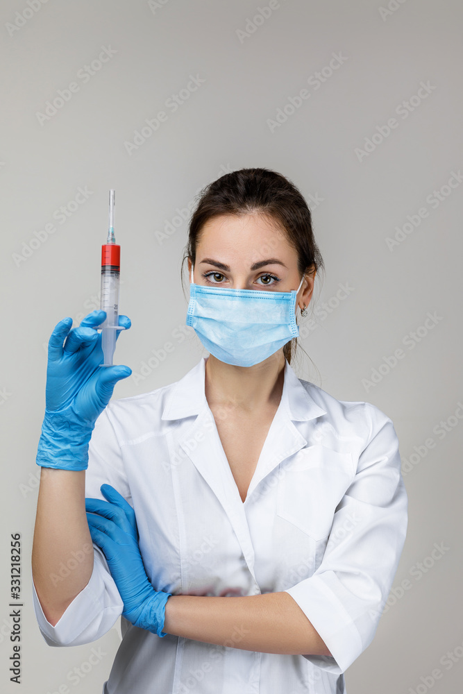 Young woman doctor in white uniform, mask and rubber gloves holding syringe on gray background. Young woman doctor in white uniform, mask and rubber gloves holding syringe on gray background.