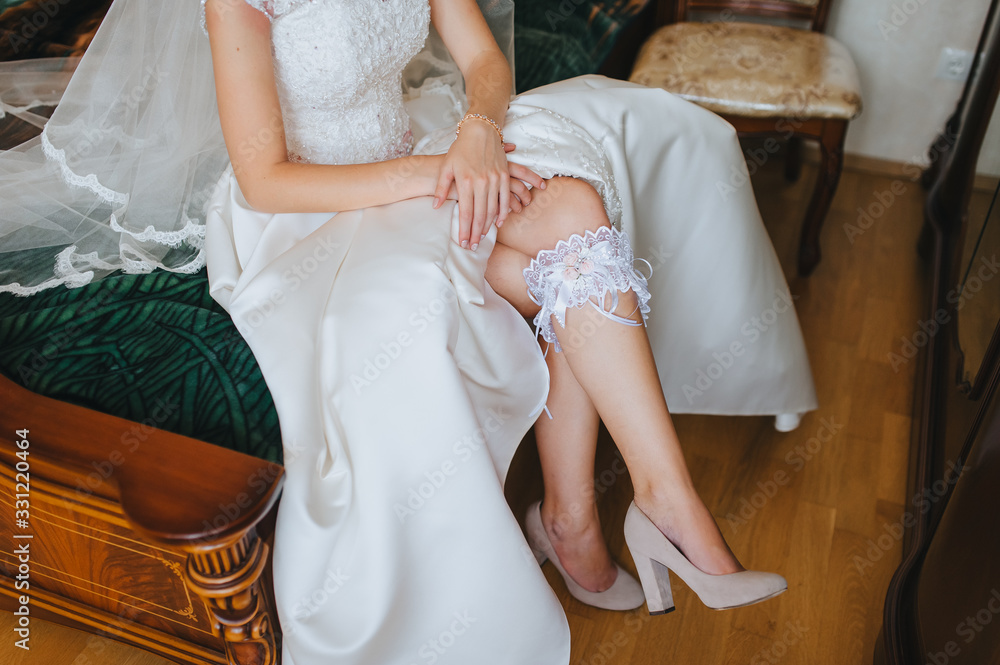 The bride sits on a sofa in a white dress with a garter on her leg. Morning preparation for the wedding. Photography, concept.