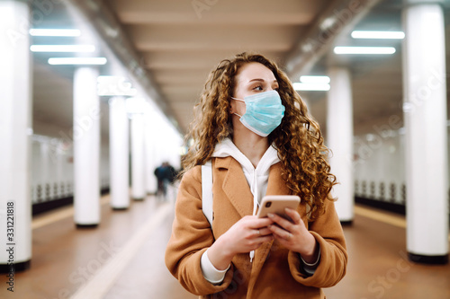 Girl in protective sterile medical mask with a phone  at subway station. Woman, wear face mask, protect from infection of virus, pandemic, outbreak and epidemic of dise ase in quarantine city.  photo