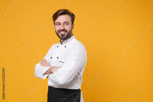 Smiling young bearded male chef cook or baker man in white uniform shirt posing isolated on yellow background in studio. Cooking food concept. Mock up copy space. Holding hands crossed, looking aside.
