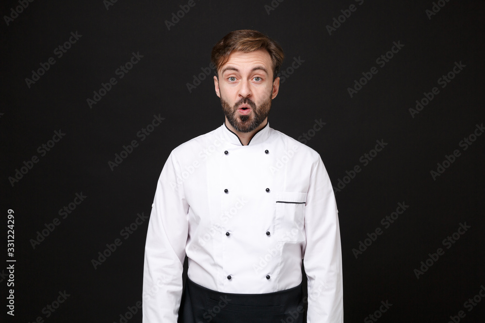 Shocked amazed young bearded male chef cook or baker man in white uniform shirt posing isolated on black wall background studio portrait. Cooking food concept. Mock up copy space. Looking camera. Shocked amazed young bearded male chef cook or baker man in white uniform shirt posing isolated on black wall background studio portrait. Cooking food concept. Mock up copy space. Looking camera.