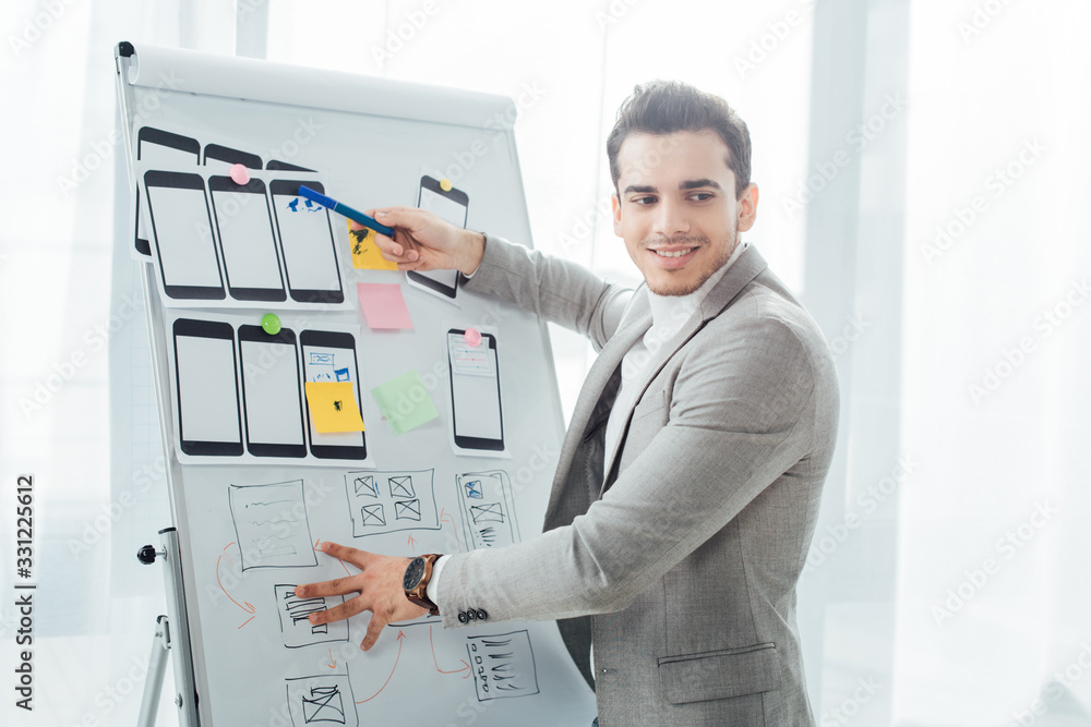Smiling ux designer pointing with marker on website sketches and mobile templates on whiteboard in office