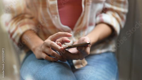 Cropped image of beautiful woman using/holding smartphone in her hands while sitting at the working desk over modern living room as background.