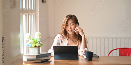 Valokuvatapetti Photo a beautiful woman using a smartphone to calling someone while sitting in front a computer tablet with keyboard case at wooden working table over comfortable living room as background