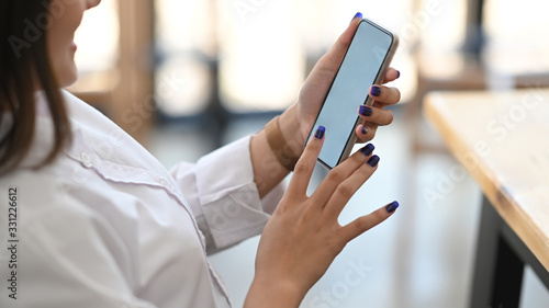 Photo of beautiful woman holding a smartphone in her hands while sitting and lean on chair at the wooden table over office glass wall as background.