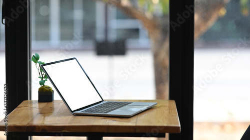 Photo of white blank screen computer laptop putting on wooden working desk with potted plant over modern office glass wall as background.