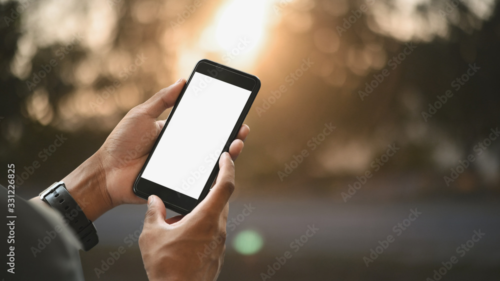 Cropped image of smart man's hands holding a cropped black smartphone with white blank screen over blurred sunny outdoors as background.