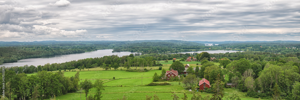 Summer green field and meadows on the hills panorama. Blue lake on the side and scandinavian traditional red wooden houses with blue sky Summer green field and meadows on the hills panorama. Blue lake on the side and scandinavian traditional red wooden houses with blue sky