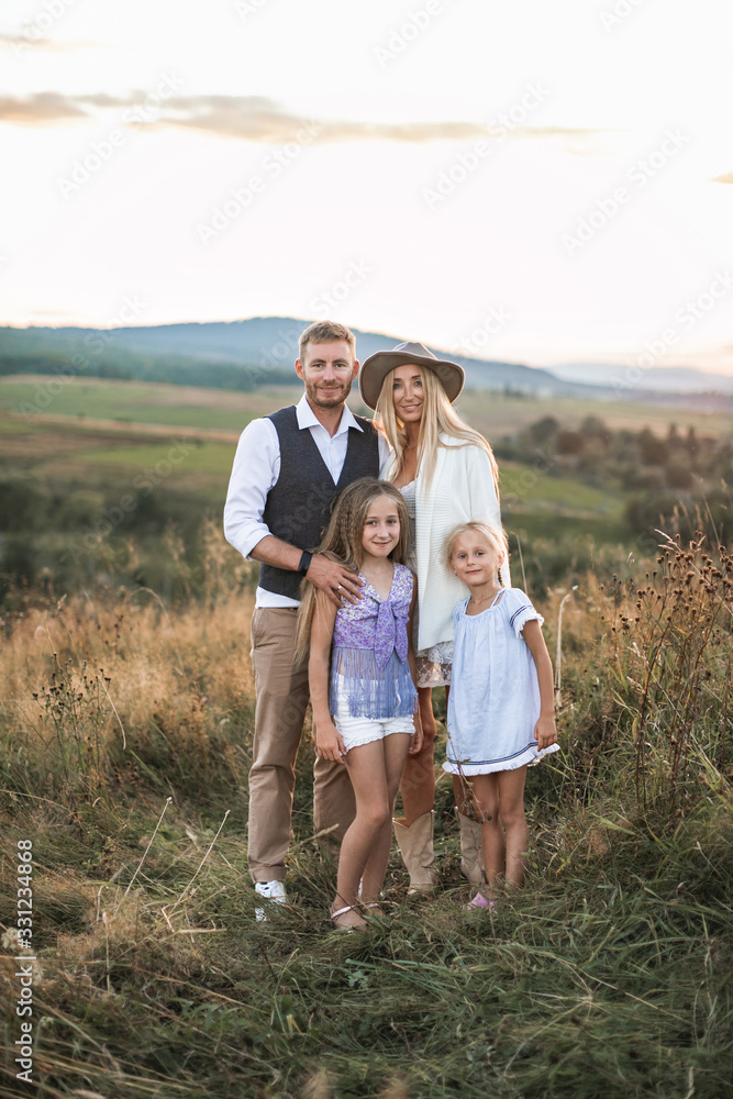 Full length portrait of cheerful Caucasian family, mother, father and two little daughters, standing in summer wild field, mountains on the background Full length portrait of cheerful Caucasian family, mother, father and two little daughters, standing in summer wild field, mountains on the background