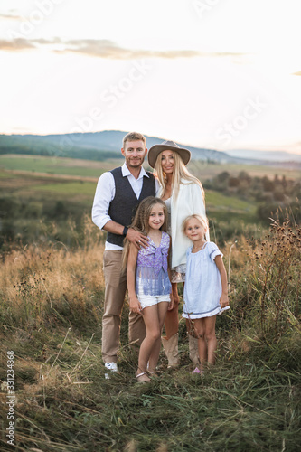 Full length portrait of cheerful Caucasian family, mother, father and two little daughters, standing in summer wild field, mountains on the background