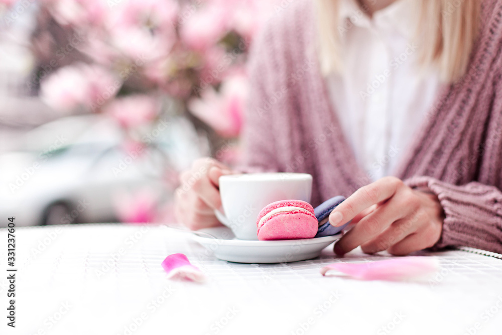 Spring cafe. Young woman drinking coffee with pink macaroons at city streets. Girl enjoying tenderness and morning breakfast outdoor. Blooming magnolia flowers. Close up of female hands. Copy space Spring cafe. Young woman drinking coffee with pink macaroons at city streets. Girl enjoying tenderness and morning breakfast outdoor. Blooming magnolia flowers. Close up of female hands. Copy space