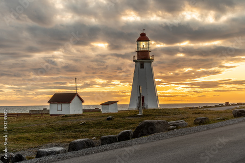 Light is shining at Point Riche Lighthouse as the sun is setting into the Gulf of St Lawrence under a cloudy sky, Port au Choix, Newfoundland, Canada